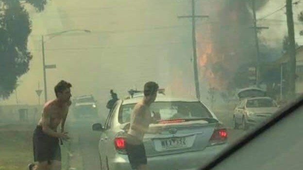 Two men with their shirts off run across a road as a fire burns behind them sending up a black plume of smoke.
