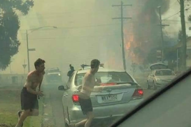 Two men with their shirts off run across a road as a fire burns behind them sending up a black plume of smoke.