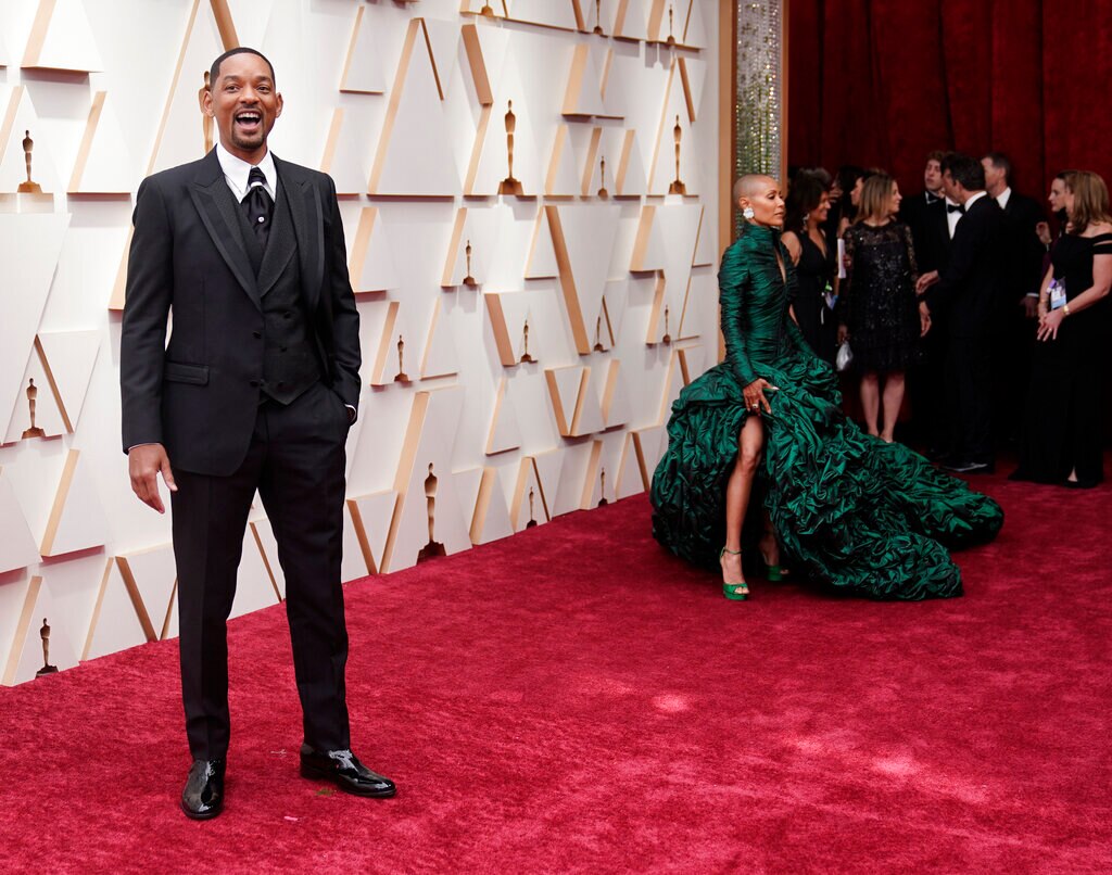 Man (Will Smith) stands on red carpet facing camera. A woman in green dress stands behind him looking away. 