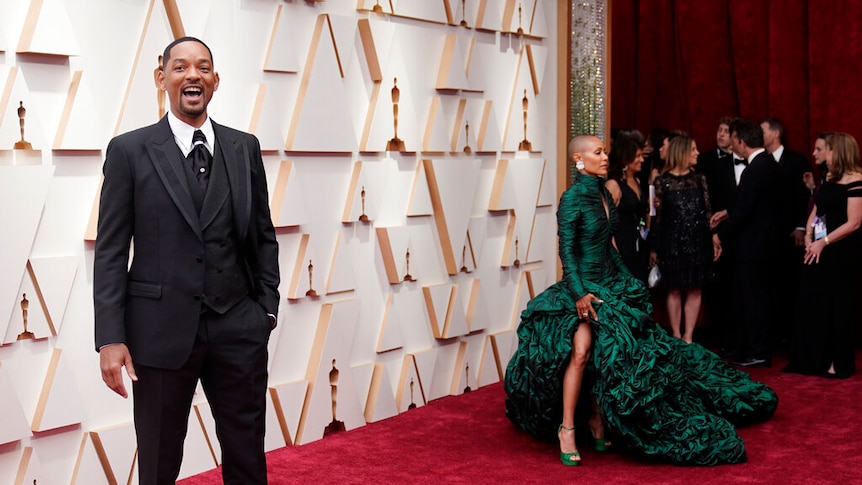 Man (Will Smith) stands on red carpet facing camera. A woman in green dress stands behind him looking away. 