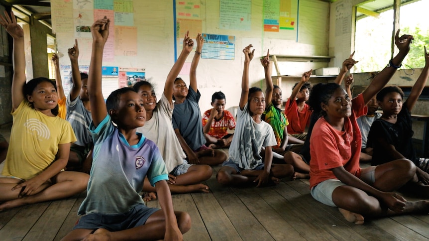 A group of children sitting on the floor of a classroom in PNG all raise their hands, looking at a teacher.