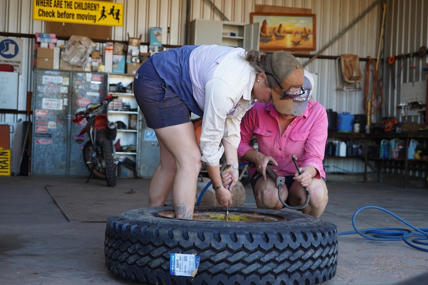 A woman in light pink shirt and blue shorts, cap, repairs a wheel in a shed while another woman in a dark pink shirt looks on.