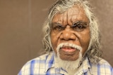 An Aboriginal man in a checkered blue and white shirt standing in front of a brown background with white hair and a white beard