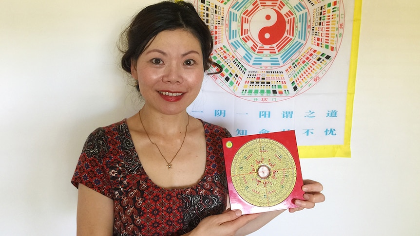 Mina Zheng standing in front of a multi-coloured Chinese astrology chart, holding a red feng shui compass.
