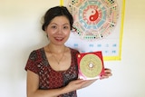 Mina Zheng standing in front of a multi-coloured Chinese astrology chart, holding a red feng shui compass.
