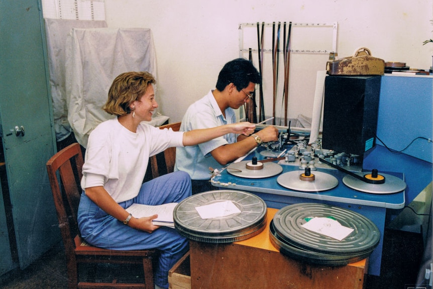 Hutcheon and film editor sitting at editing machine with spools of film threaded through machine.