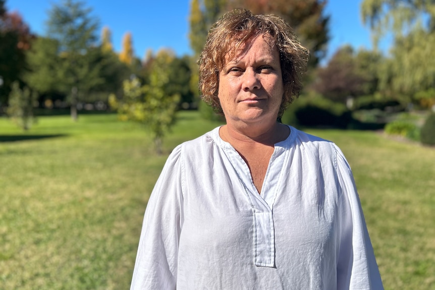 An Indigenous woman in a white collared shirt stands in a field looking serious.