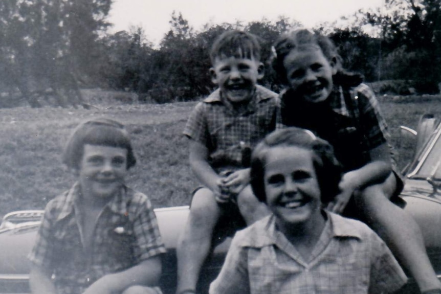 A black and white image of four children sitting on the front of a car in the 1950s.
