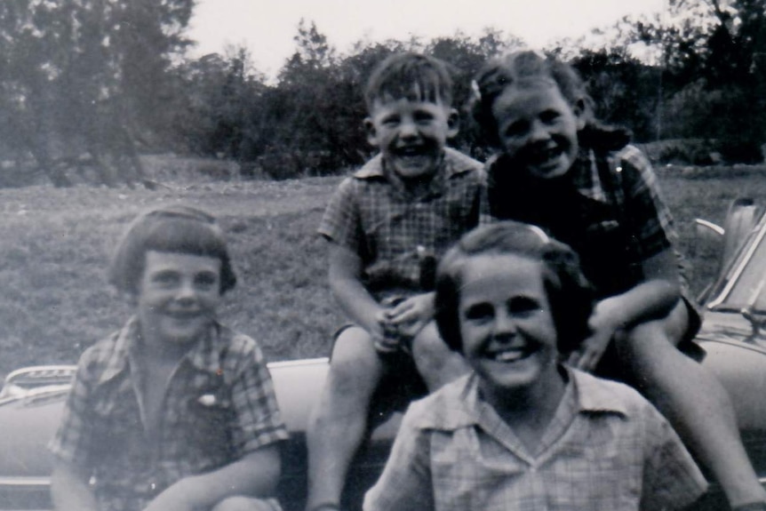 A black and white image of four children sitting in front of a 1950s car.