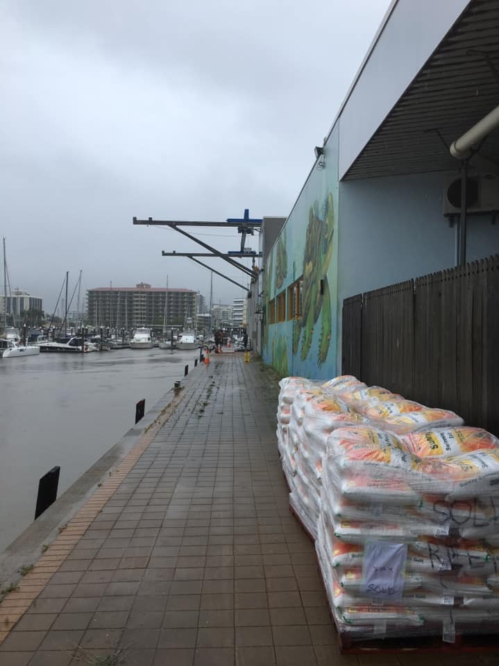 Four pallets of pool salt sit on the exterior wall of a building next to a marina filled with boats