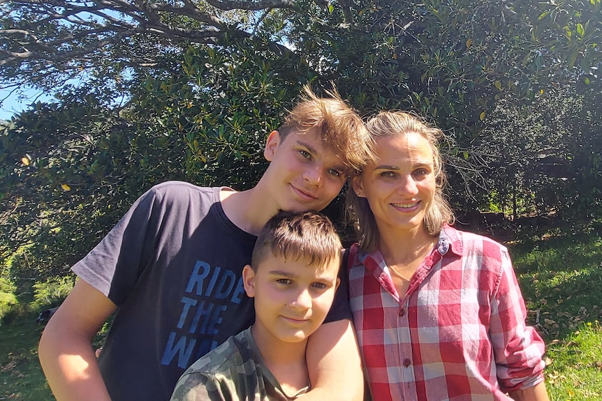 Mother and two sons hug and smile in front of large tree 