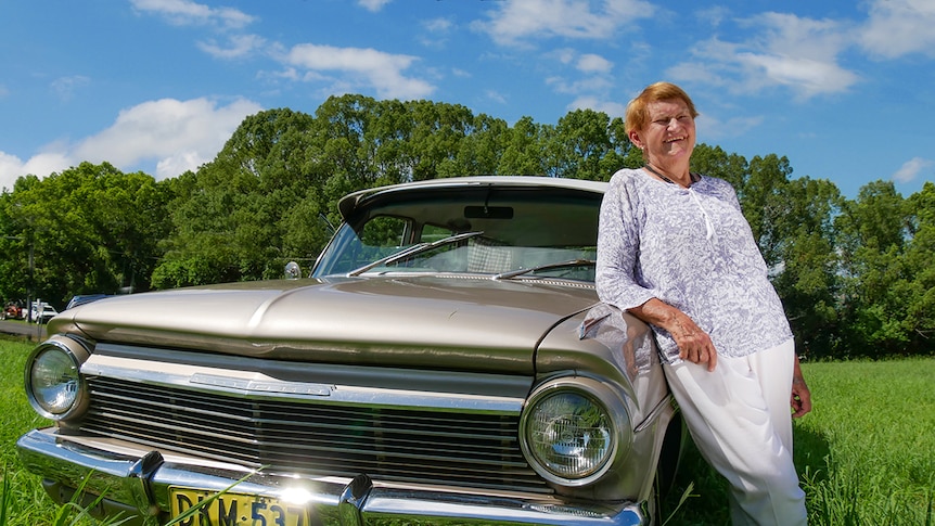 Woman reclines on her shiny holden car bonnet surrounded by green trees