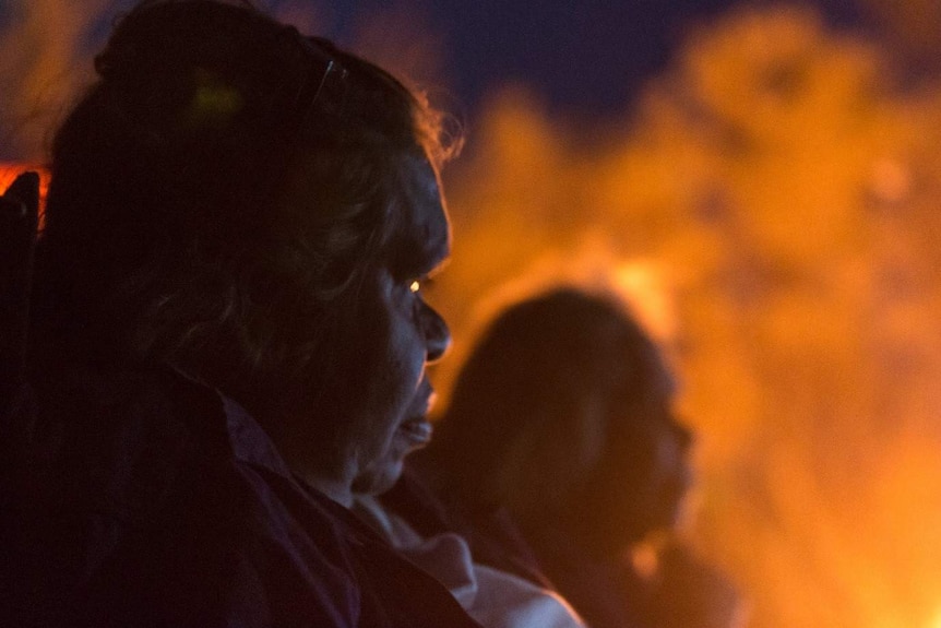 An Indigenous woman's face is lit in profile by a campfire.