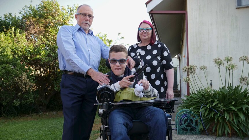 A boy in his wheelchair is surrounded by his two parents in a portrait in their backyard