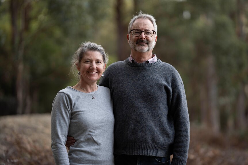 A man and woman stand together smiling with trees in the background