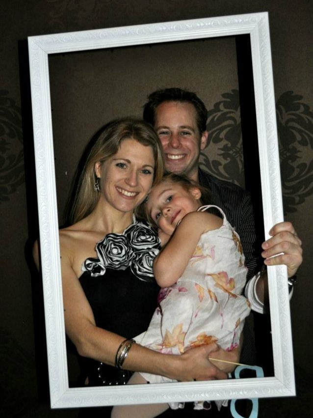 A happy couple dressed up in formal wear hold a smiling girl in their arms.