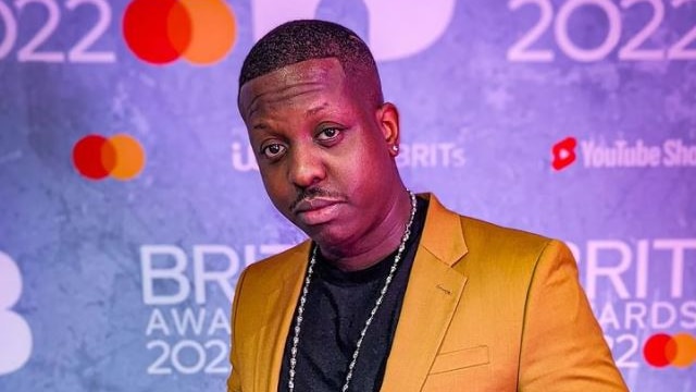 Jamal Edwards wearing a yellow suit in front of the Brit Awards 2022 backdrop. 