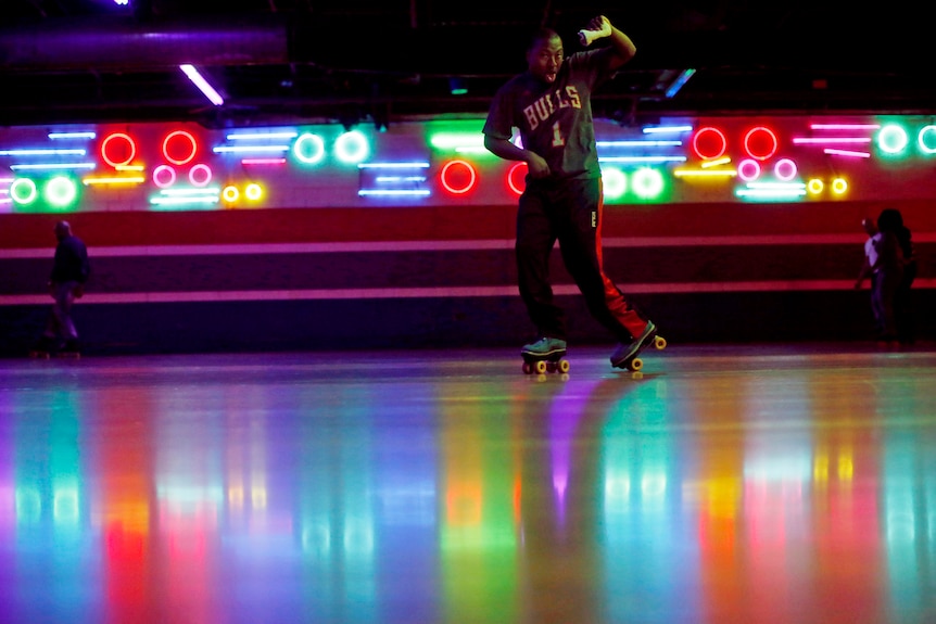 A man skates at a rink with retro fluorescent lights and colours flashing behind him 