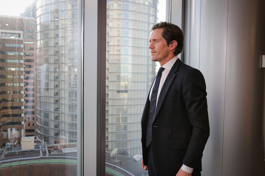 A slender man in a dark suit and patterned tie stands side-on, beside a picture window overlooking other high-rise offices.