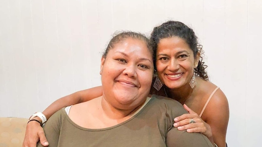 A woman sitting and another woman smiling and wrapping her arms around her.