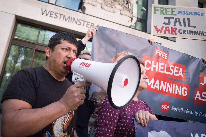 Man speaks into a megaphone in front of signs calling for Julian Assange to be freed.