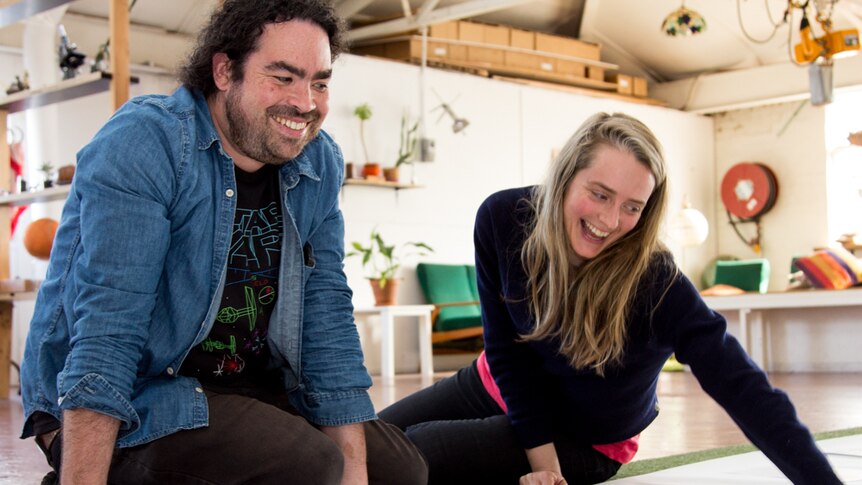 Two people sit on the floor with drawings spread out in front of them.