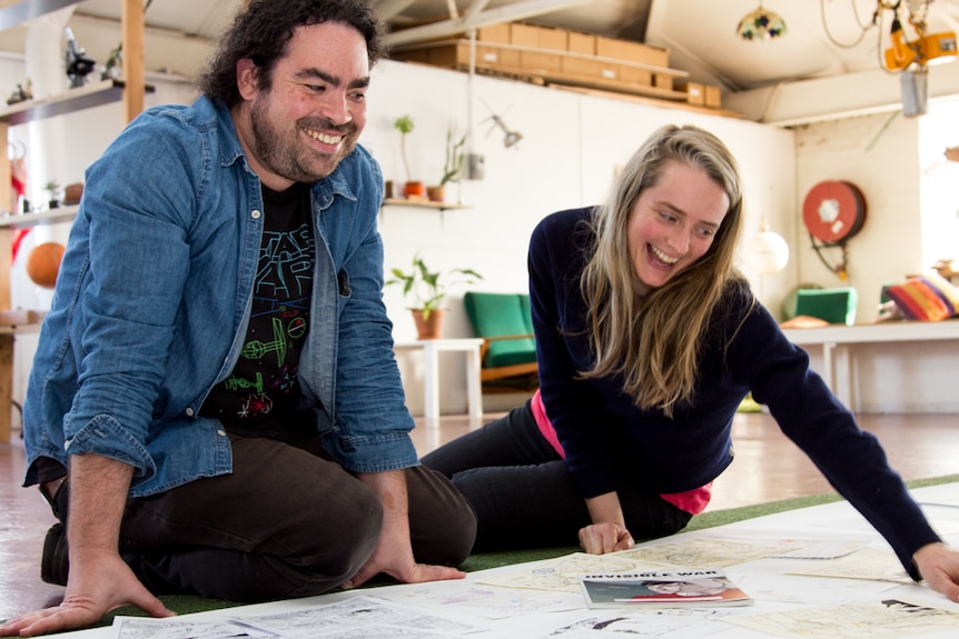 Two people sit on the floor with drawings spread out in front of them.