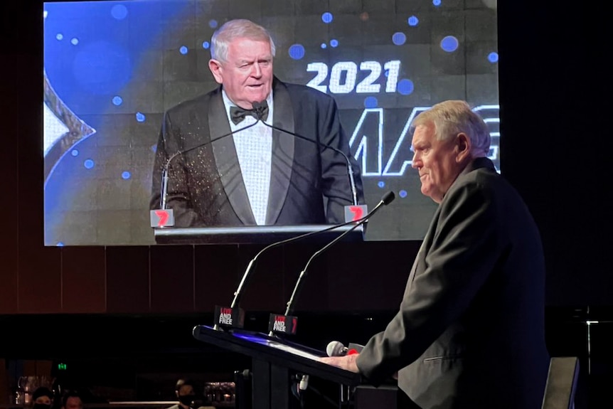 A man in a tuxedo stands at a podium with his image on a big screen in the background.