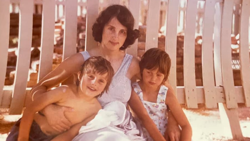 A woman with dark hair sits against a fence with her arms around two young children.