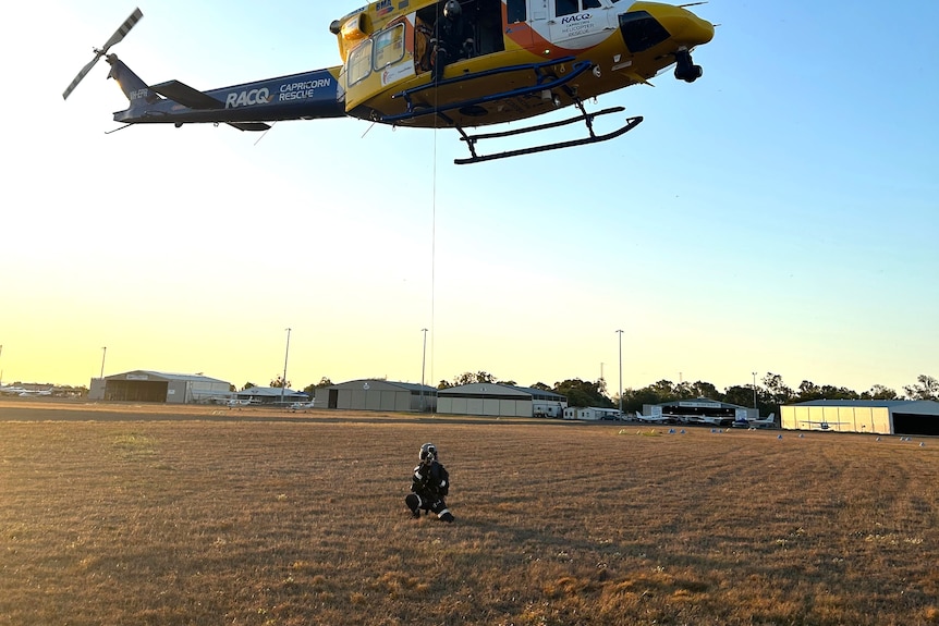 A rescue crew officer lands on the ground of a helicopter which is yellow, white and blue