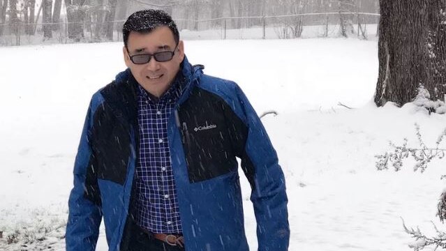 A mid-age Chinese man wearing a down jacket standing outdoor while it was snowing.