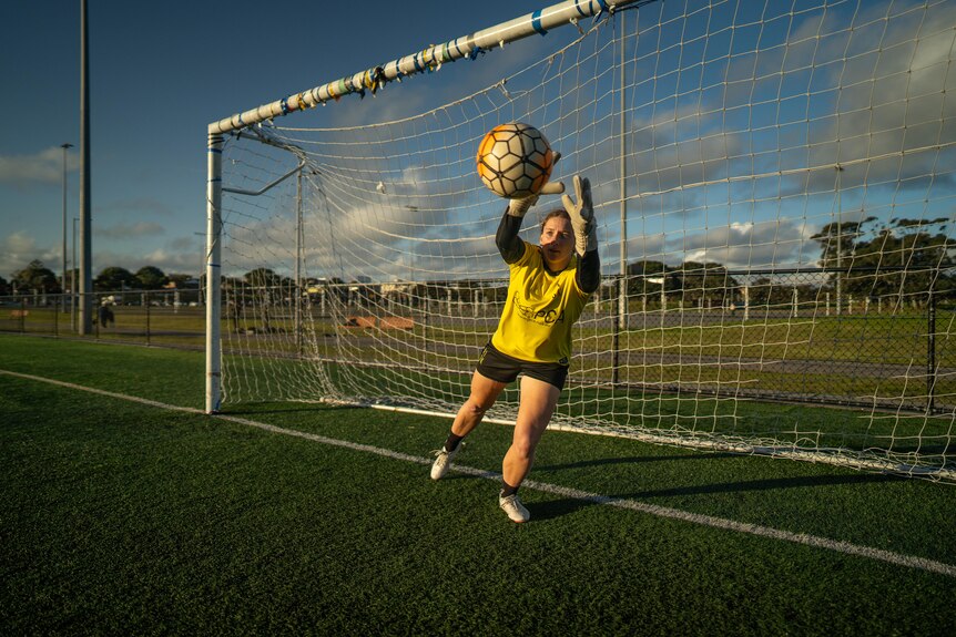 Female soccer goalkeeper in yellow jersey and shorts catching a soccer ball at sunset.