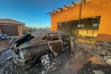 A burnt out car in front of a damaged building are shown at dawn in bright sunlight
