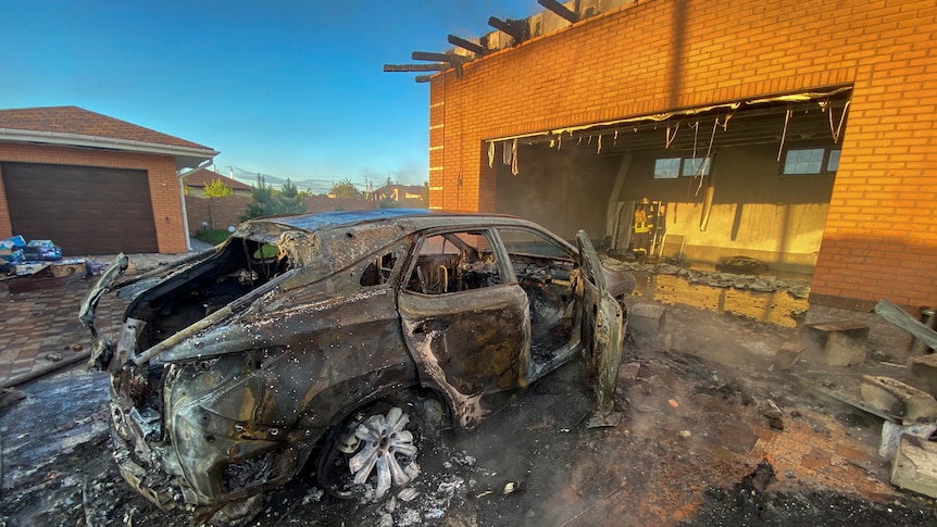 A burnt out car in front of a damaged building are shown at dawn in bright sunlight