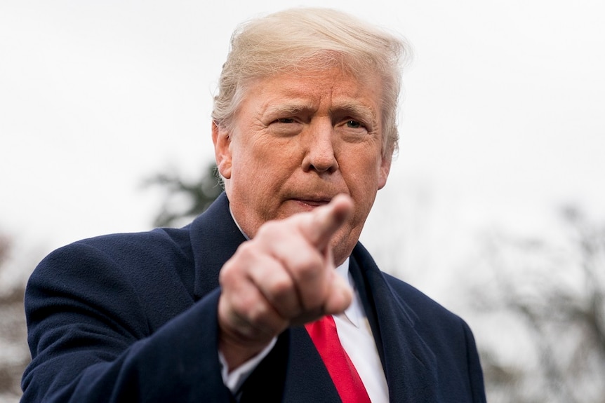 A man in a dark coat points his right index finger as he prepares to speak to people in front of him off camera.