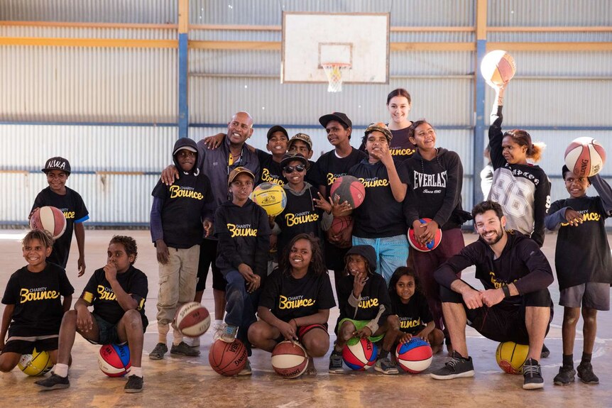 NBL star Kevin White sits on a basketball with a group of Indigenous youth on a basketball court.