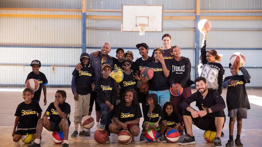 NBL star Kevin White sits on a basketball with a group of Indigenous youth on a basketball court.