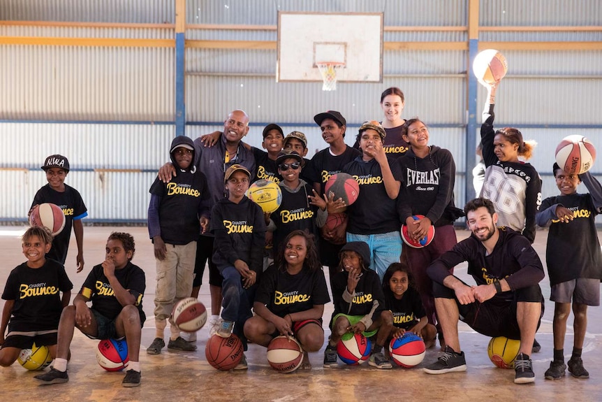 NBL star Kevin White sits on a basketball with a group of Indigenous youth on a basketball court.