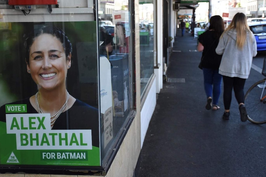 A green sign with Alex Bhathal's face on it sits in a shop window. People walk past on the street.