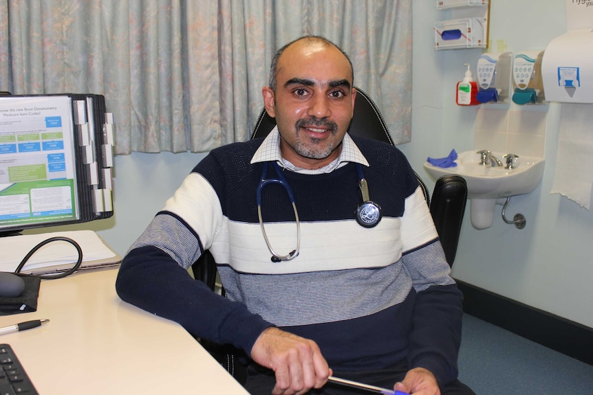 A male doctor, wearing a stethoscope, sits at his desk