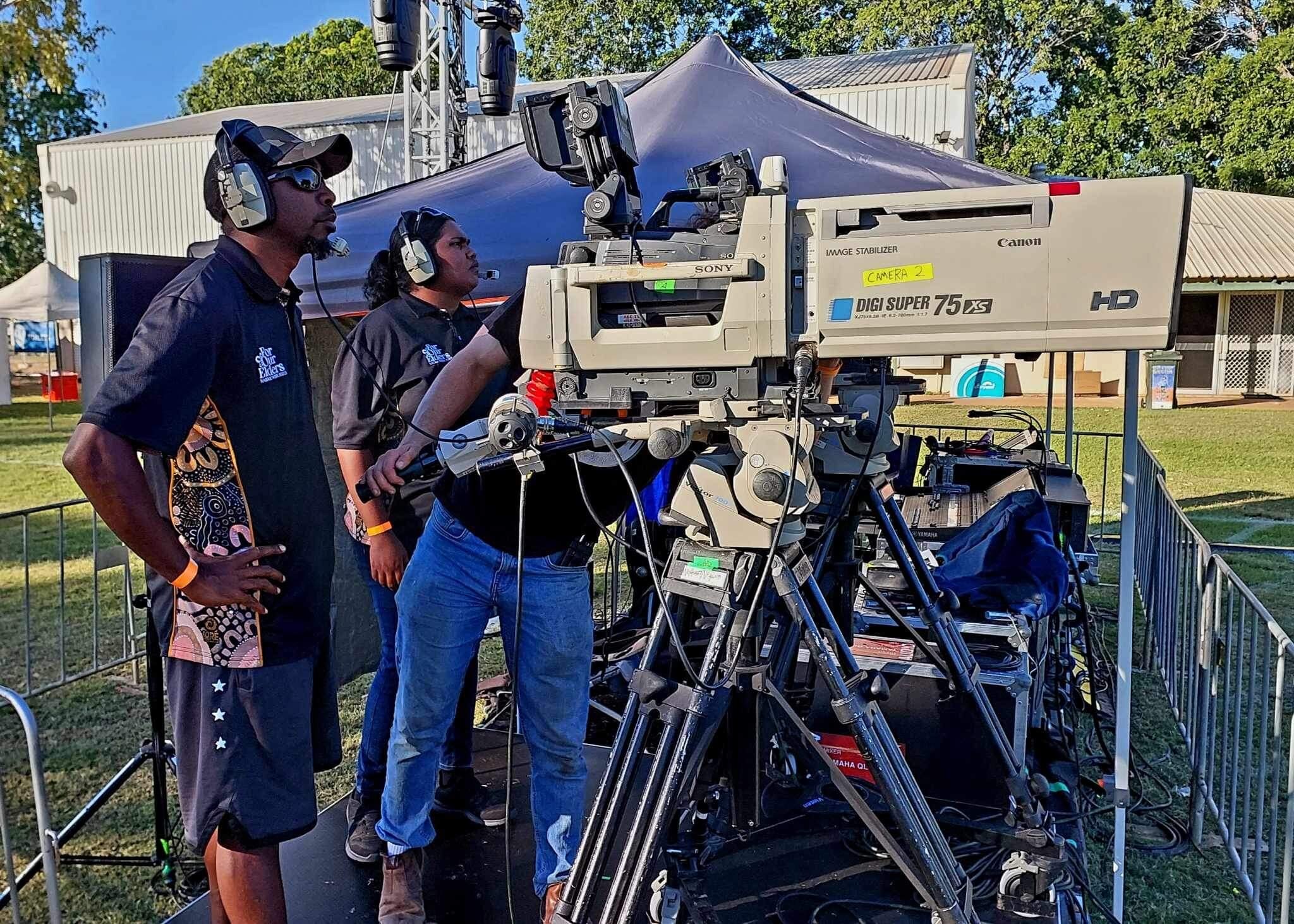 Two people wearing headsets operating large broadcast camera as a man, face hidden, adjusts camera between them.