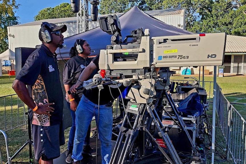 Two people wearing headsets operating large broadcast camera as a man, face hidden, adjusts camera between them.