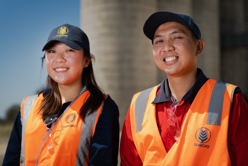 A young man and woman in high-vis vests labelled 'GrainCorp' stand smiling in front of a grain silo.