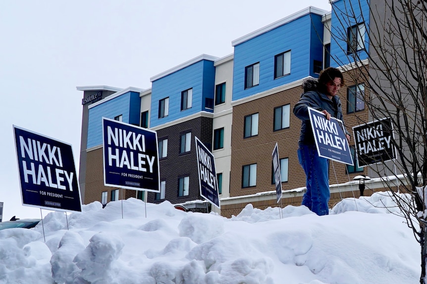 A man puts a blue sign that says 'Nikki Haley' in the snow. Other signs are already standing.
