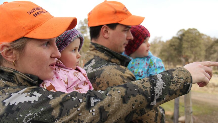 A hunting couple, dressed in camouflage but wearing orange caps, hold their children and point to wildlife