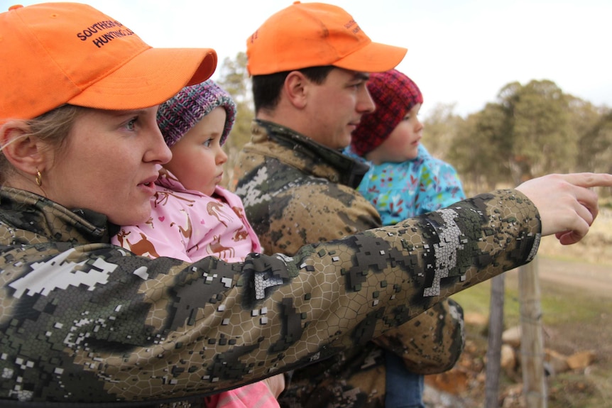 A hunting couple, dress in camouflage but wearing orange caps, hold their children and point to wildlife