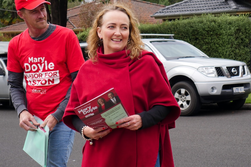 A smiling woman in a red poncho holds a stack of flyers as she crosses a road