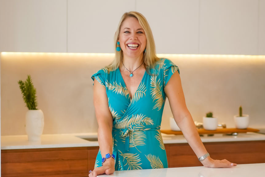 Woman in her forties with blonde hair wearing a blue dress standing in a kitchen smiling dircetly at the camera. 
