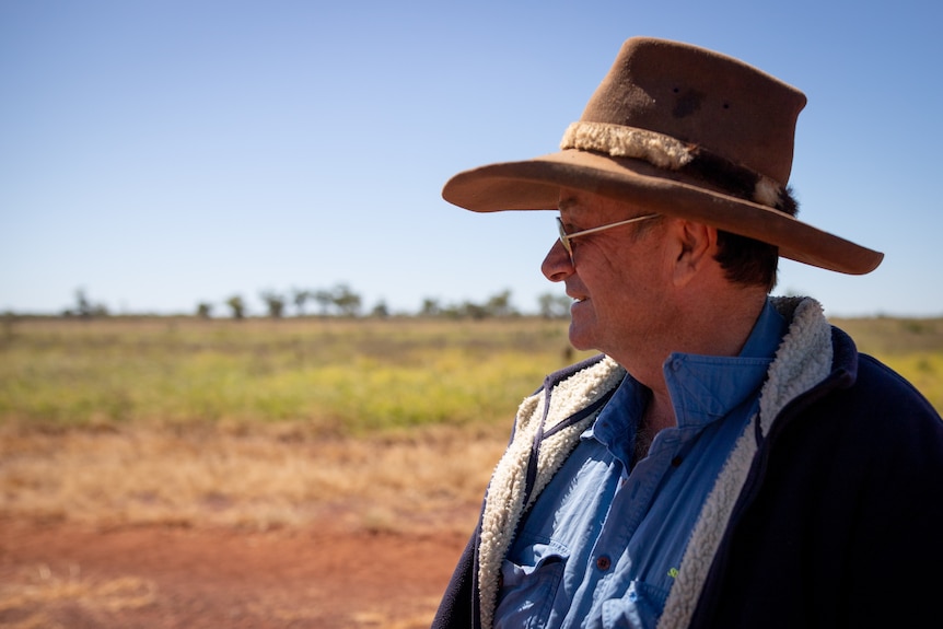 A male pastoralist wears a wide-brimmed hat and sunglasses and looks into the distance across an outback paddock. 