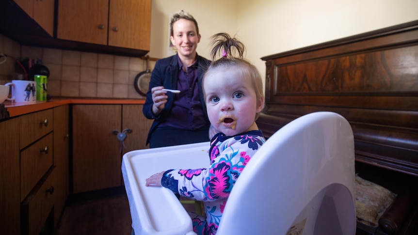 Toddler in high chair looking at camera, mother in background holding spoon and smiling